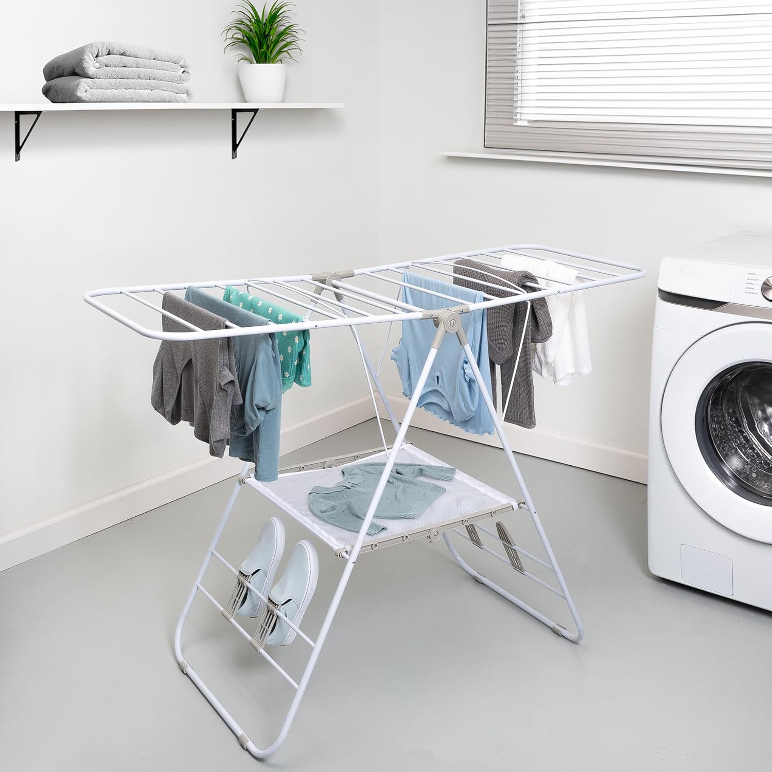 The matching white drying rack in a laundry room with garments over the rails, a garment laid on the mesh shelf and shoes on lower holders.