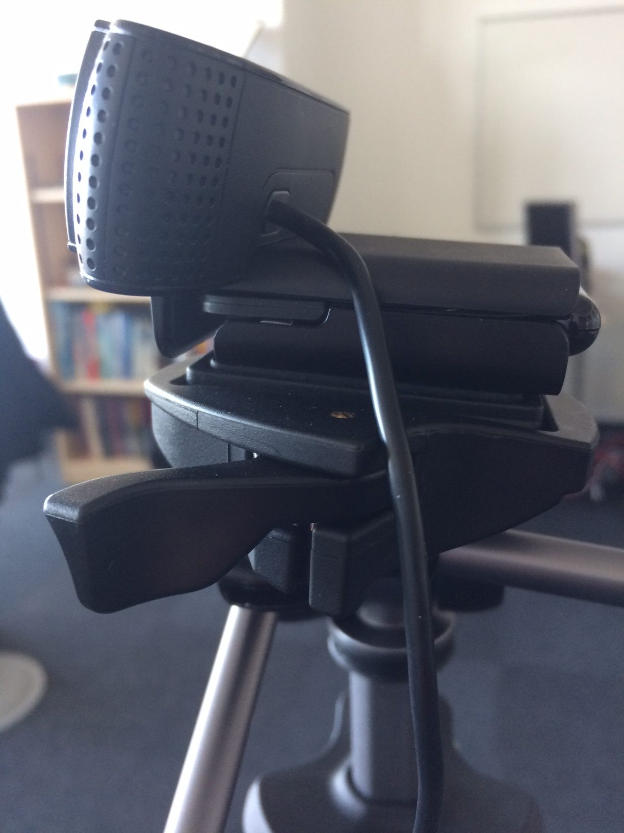 Rear side of a black Logitech C920 showing its attached cable and hinged clip on a separate tripod.