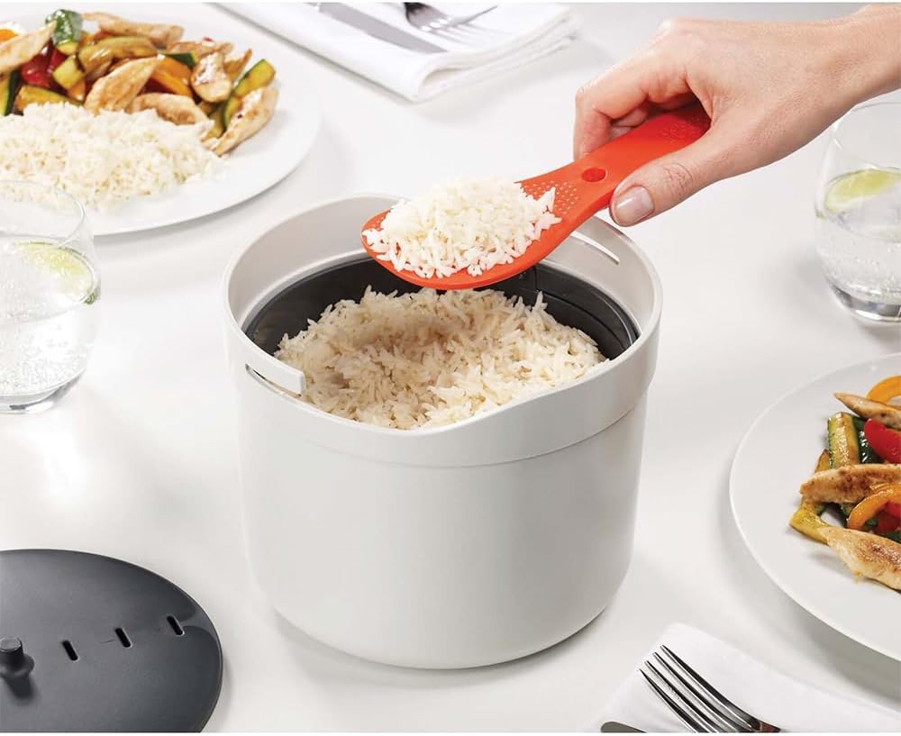 An orange paddle serves rice from the open Joseph Joseph M-Cuisine pot with its dark colander inside.