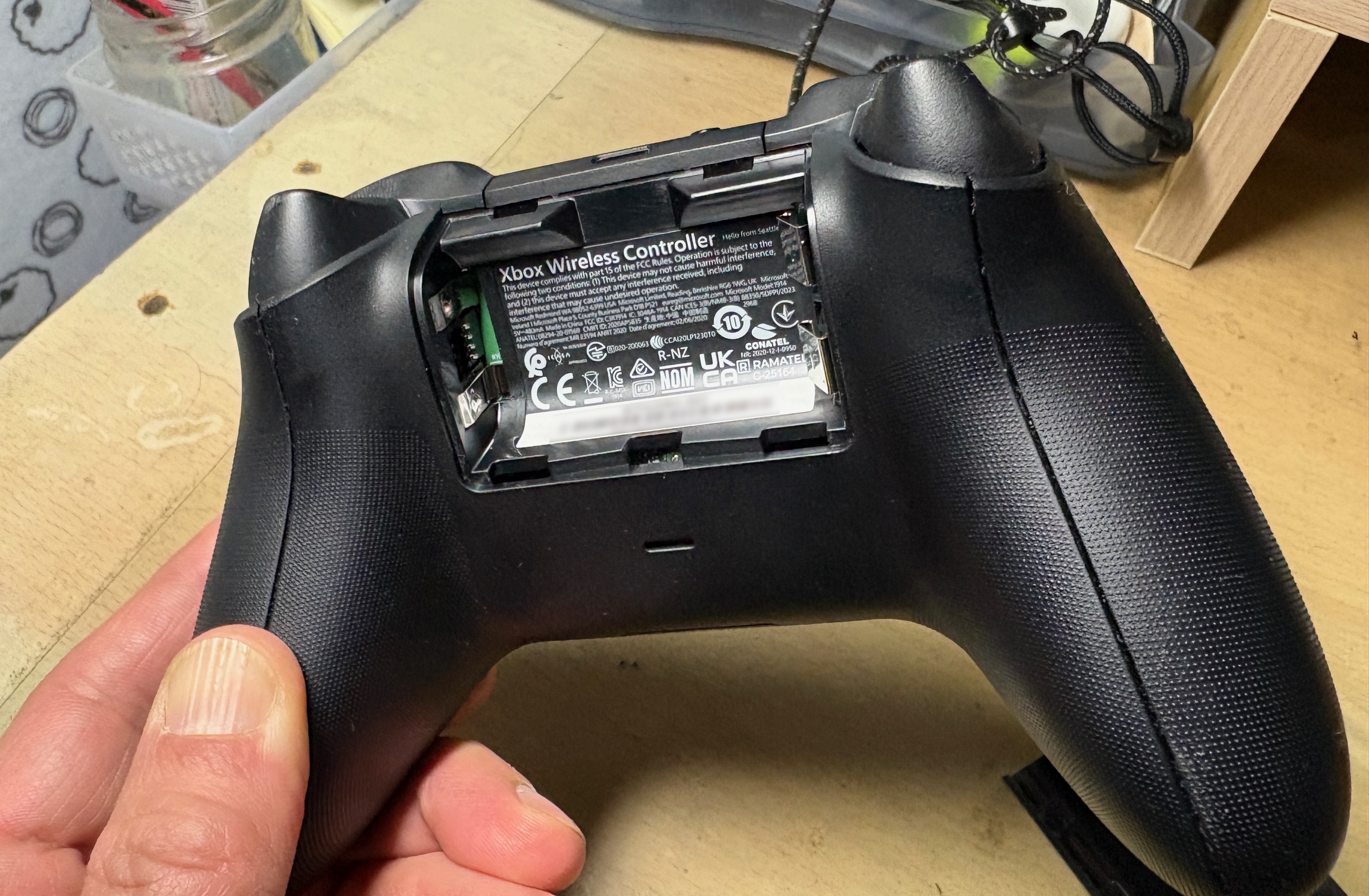 Rear of a black Xbox Wireless Controller with its battery cover removed, empty AA compartment, and textured grips.