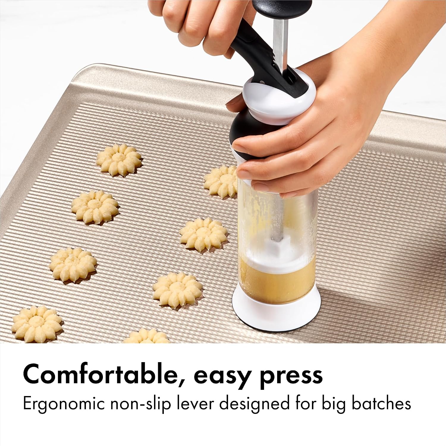 Hands operating the OXO cookie press against a metal baking sheet, with dough in the clear barrel and flower-shaped pieces on the tray.