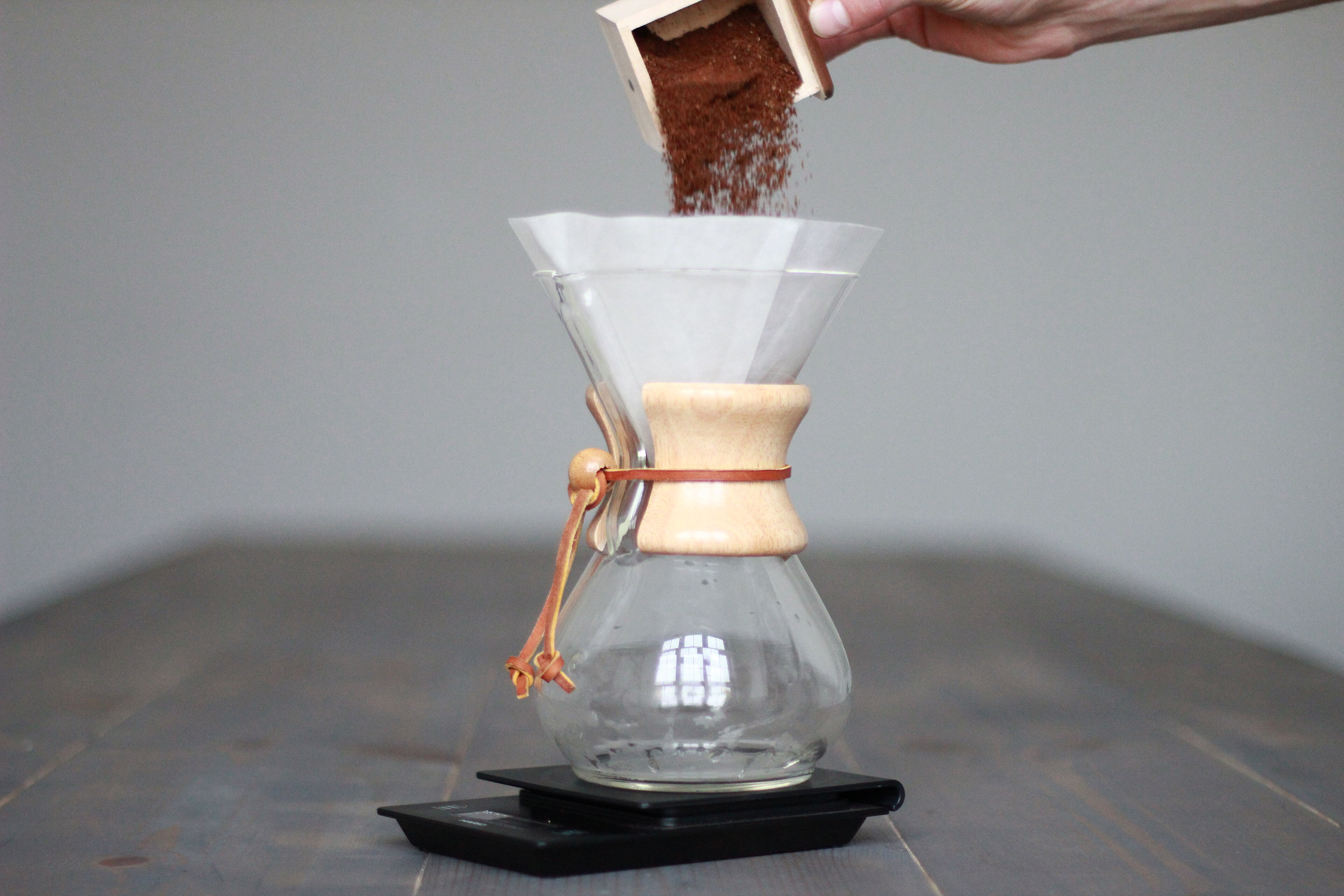 Ground coffee being added to a paper filter in a six-cup Classic Chemex resting on a black scale.