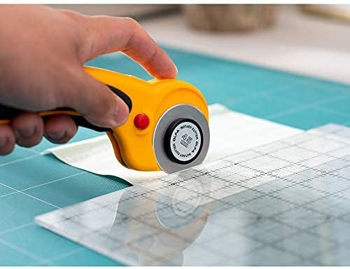 A hand holds the yellow and black OLFA RTY-2/DX beside a transparent ruler on white fabric and a turquoise cutting mat.