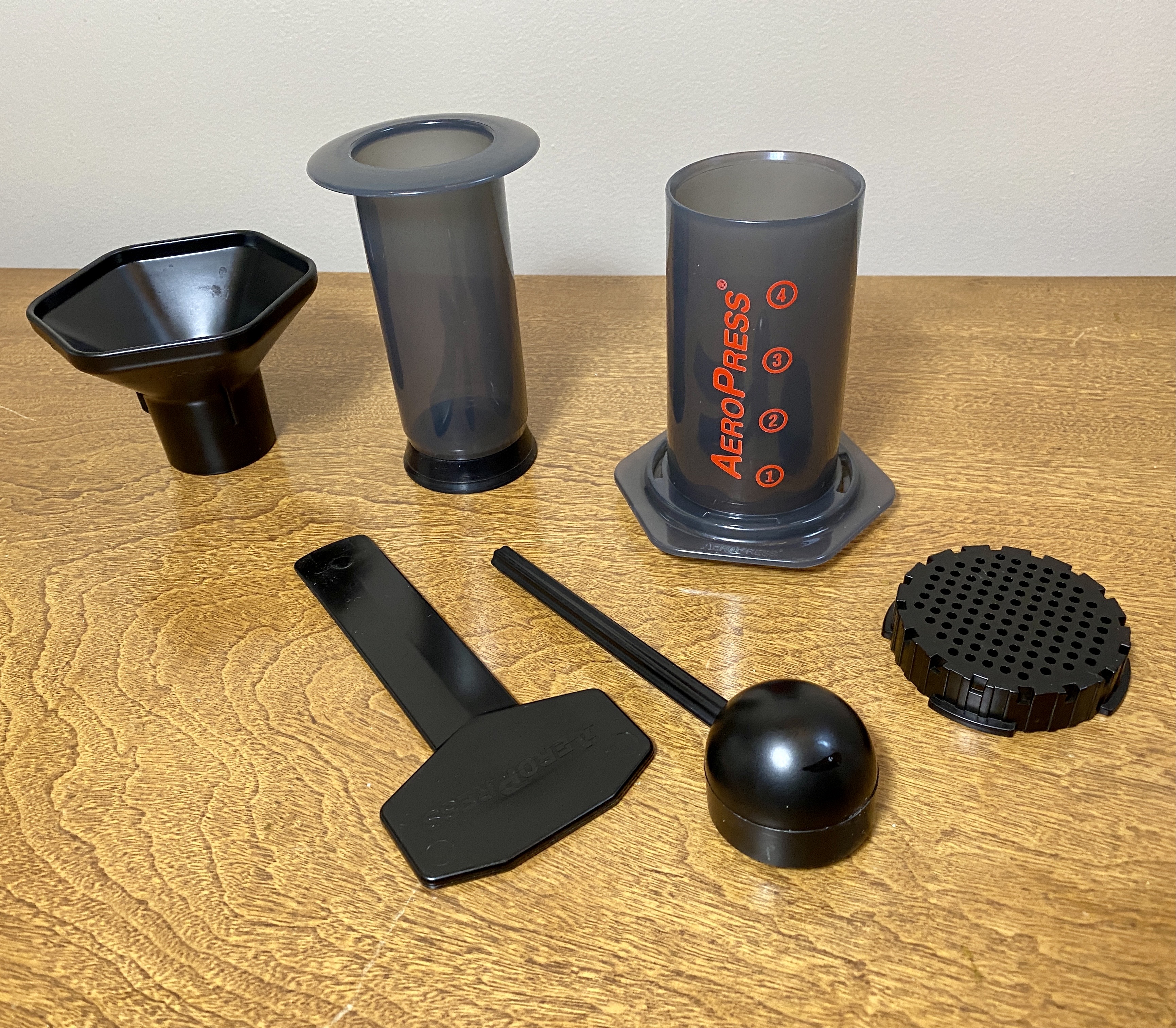 Gray AeroPress Original with red lettering, plunger, filter cap and accessories on a wooden table.