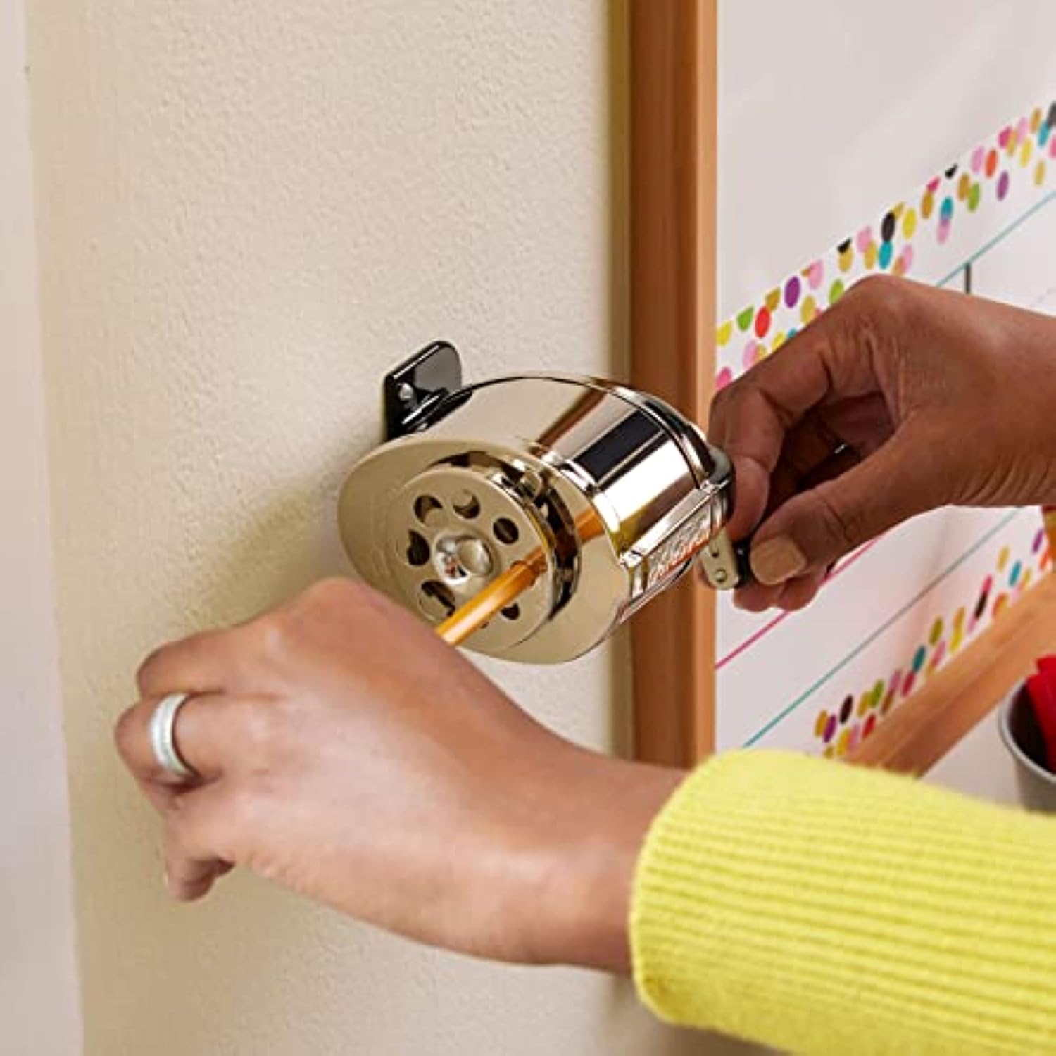 A wall-mounted X-ACTO KS sharpener with a pencil inserted into its guide, one hand supporting the pencil and another at the crank.